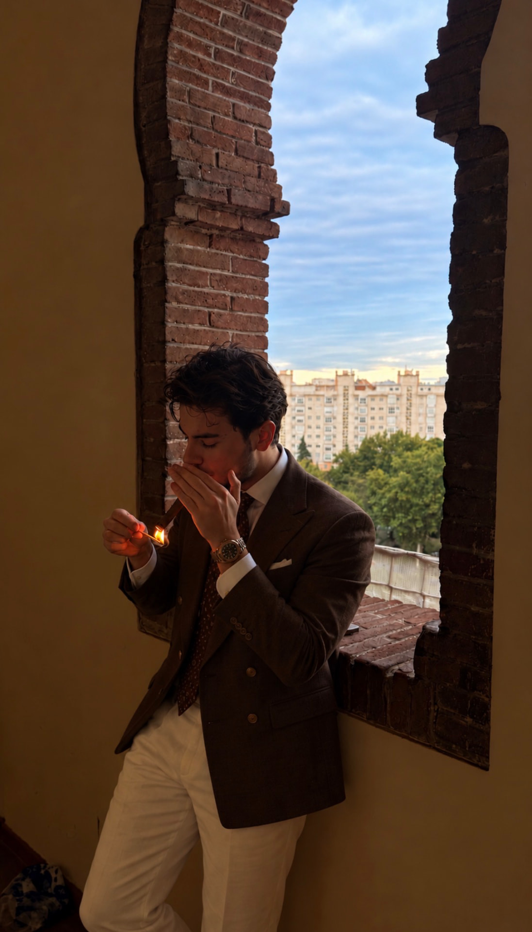 Efe Hazar Öztürk lighting a cigar by an arched brick window
