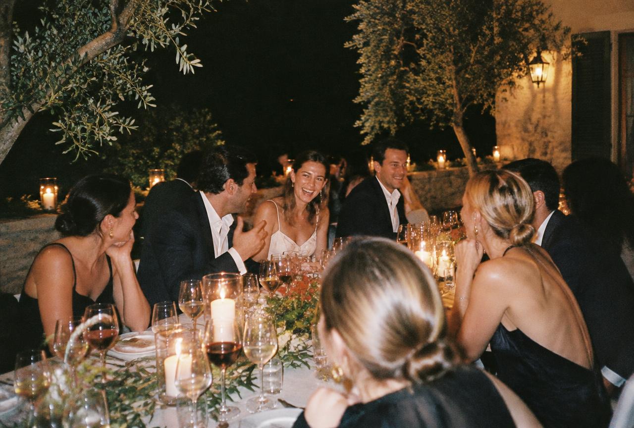 A long candlelit dinner table under olive trees